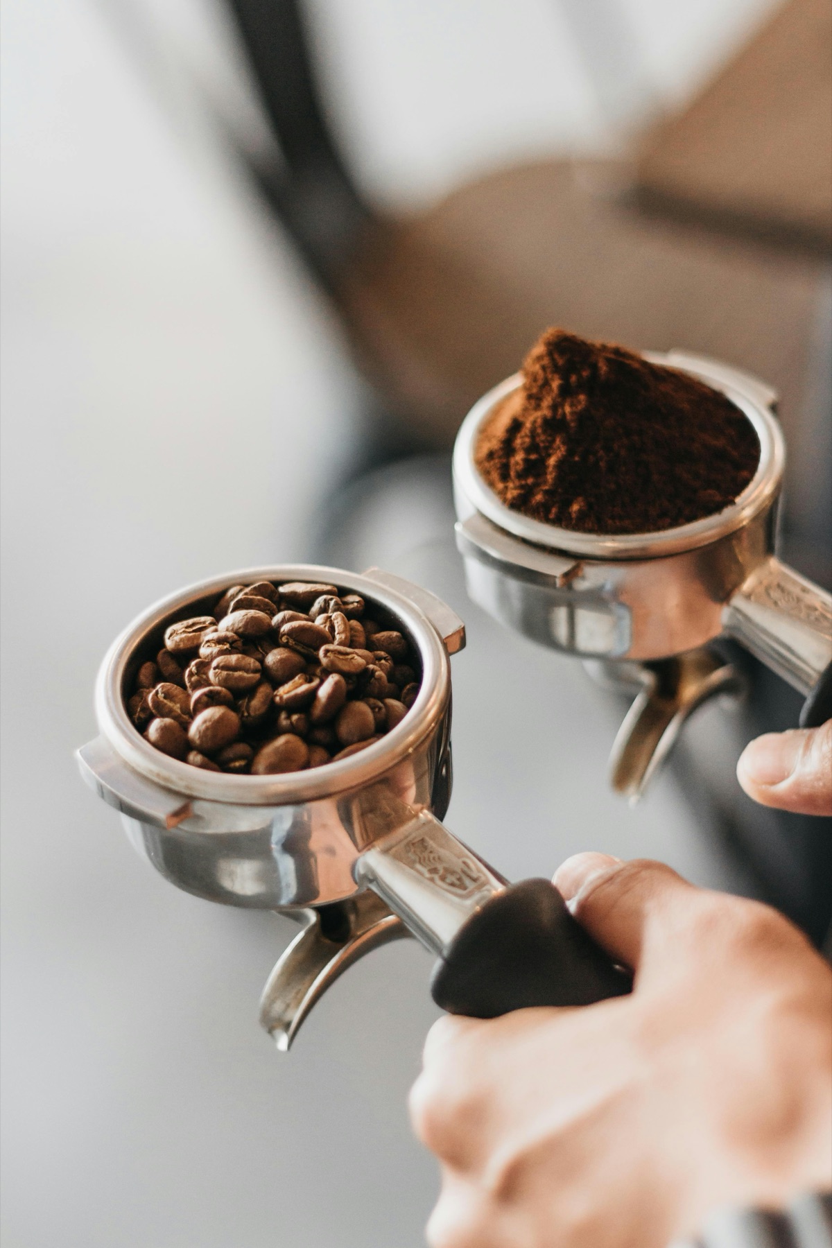 Two hands holding portafilters side by side, one filled with roasted coffee beans and the other with freshly ground coffee