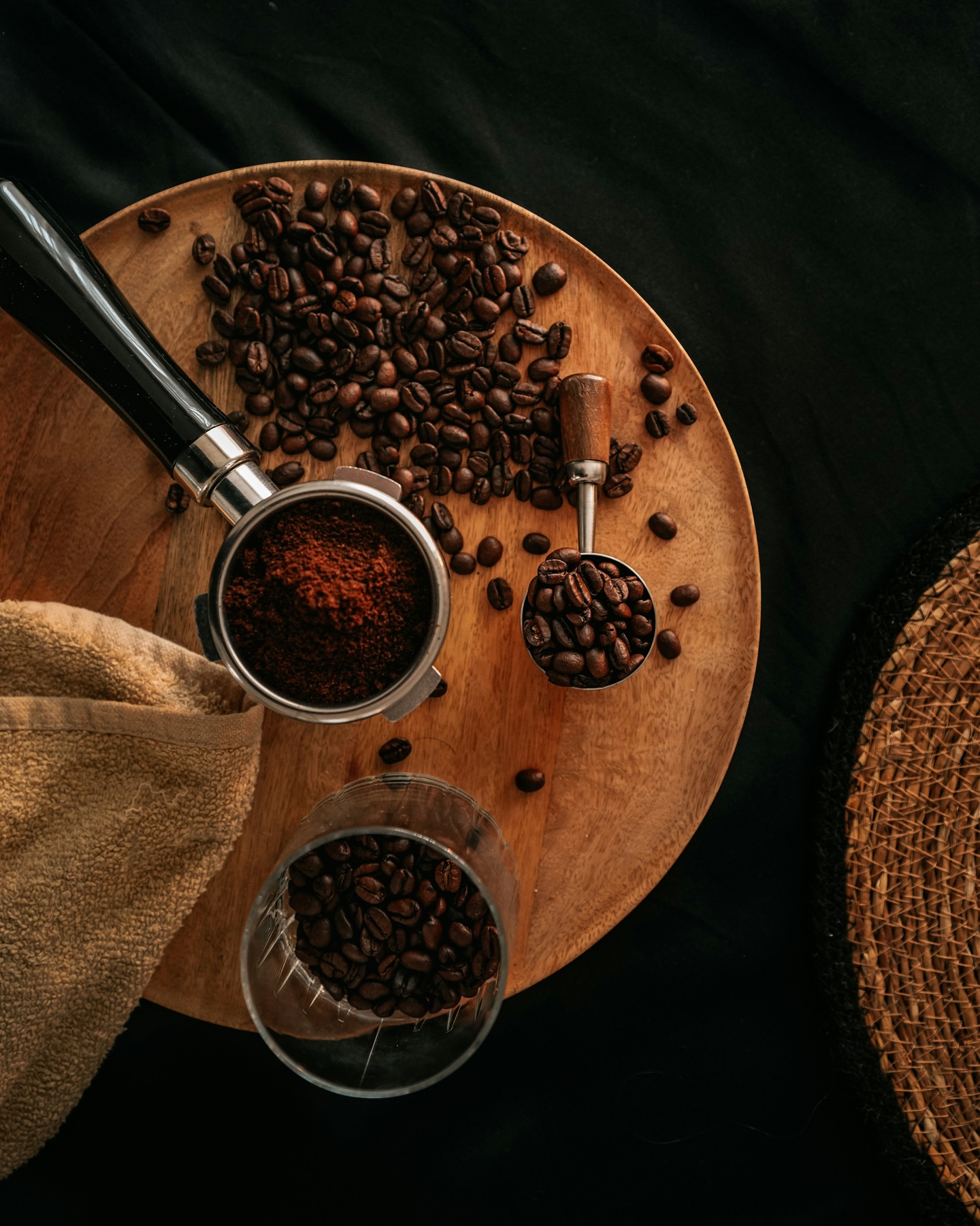 Overhead view of a round wooden board on dark cloth with a portafilter of fresh grounds, a scoop and a glass of roasted beans
