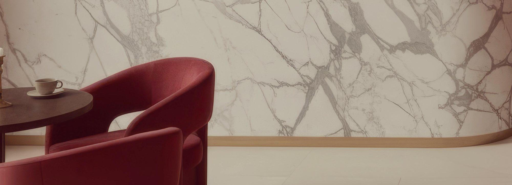 A curved wall of white veined marble with two burgundy armchairs beside a dark round side table holding a cup and saucer