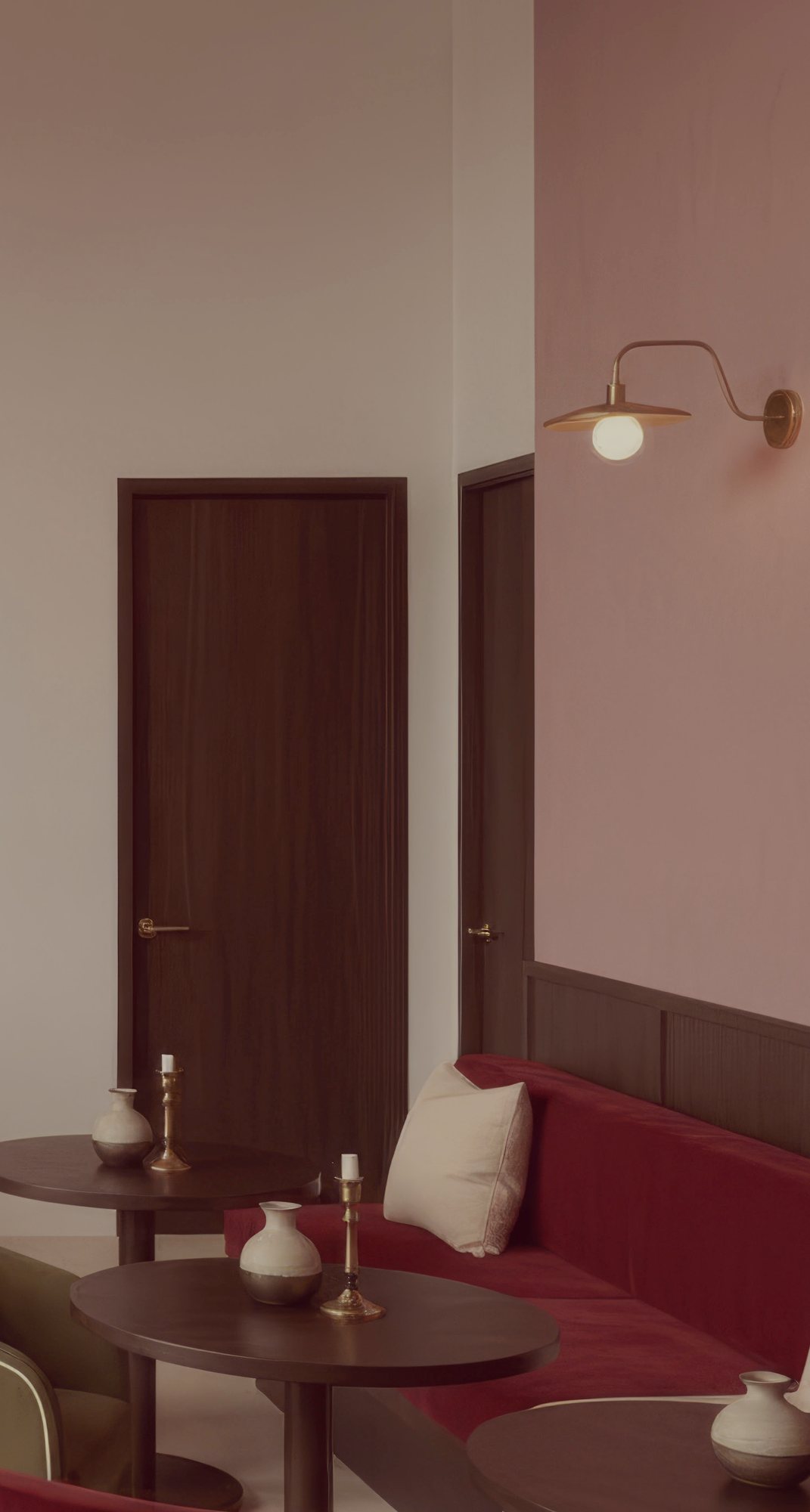 A lounge corner with a burgundy banquette, cream cushion and small round tables set with brass candlesticks and vases, under a brass wall sconce