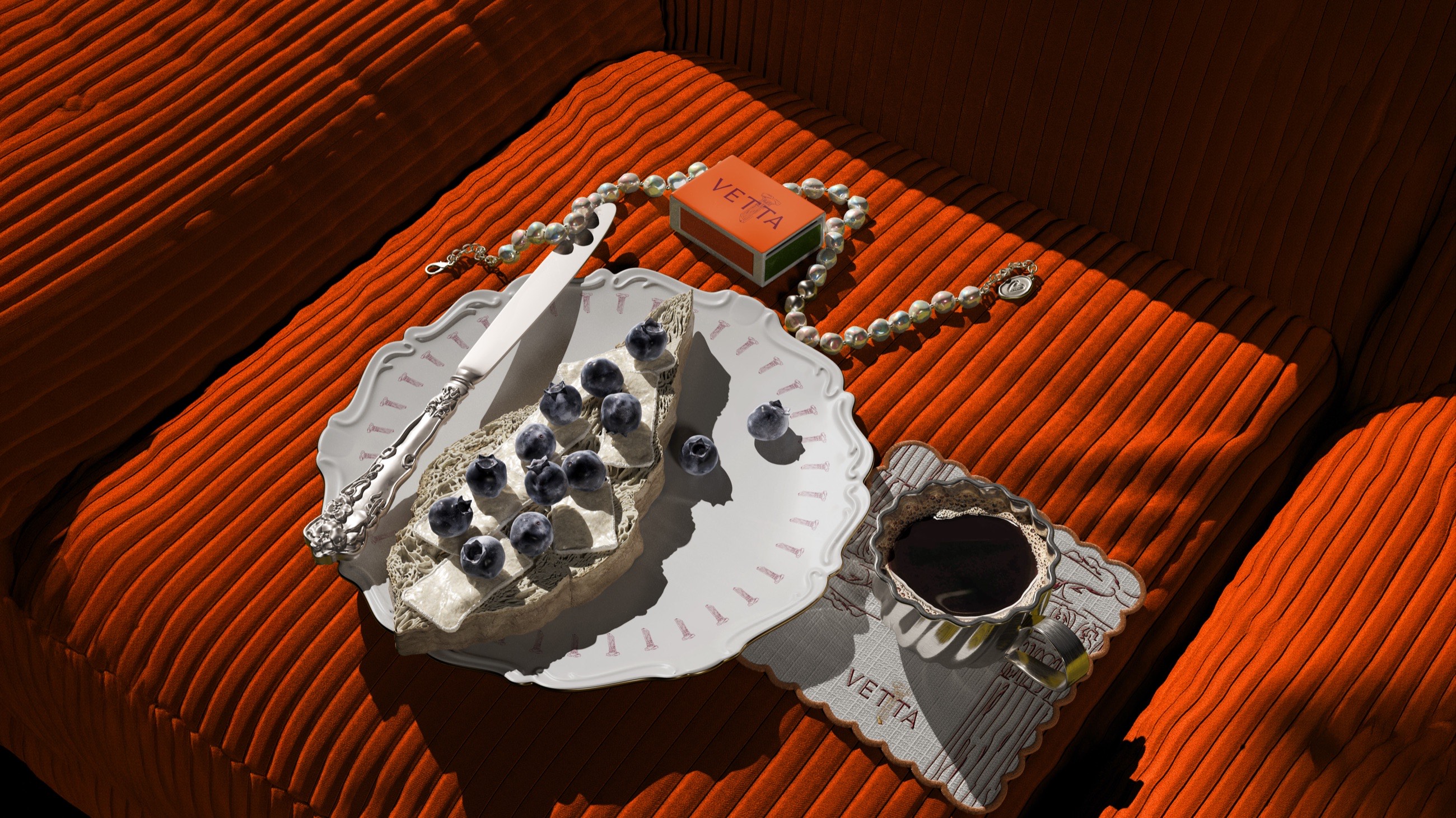 A table set on an orange corduroy cushion: a plate of bread and blueberries with a silver knife, a VETTA matchbox, a string of pearls, and a cup of black coffee on a VETTA napkin